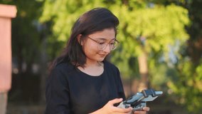 Confident talented Indian girl holding remote control controls the quadcopter drone in park during sunset. Professional aerial cinematographer taking drone shots for travel vlogs.  