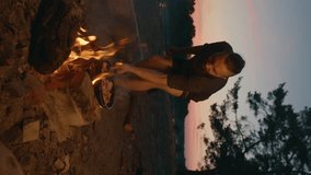 A young hipster man fries meat in a pan on a fire near the river at sunset. Slow motion. Vertical video