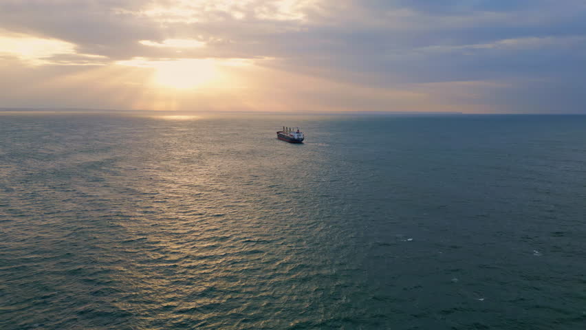 Aerial view ship sailing to picturesque sunset slow motion. Big transportation vessel floating on calm ripple surface dark ocean. Golden sunlight shining through gray clouds at dramatic summer evening