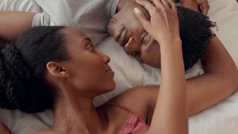 Love, couple and bed with a man and woman kissing while lying in the bedroom together in their home. Romance, happy and smile with a male and female sharing an intimate moment in the morning Video Stok