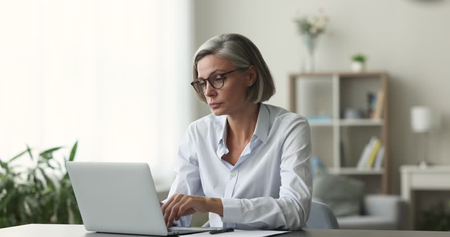 Focused middle-aged woman sit at desk, using laptop, looking pensive or concerned, learn new application, working on research project online on computer, do remote work, think about issue solution Royalty-Free Stock Footage #1102040381