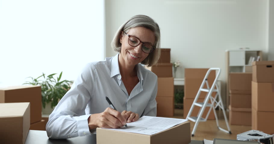 Happy small business owner prepare parcel for sending, sit at desk writes address, shipping goods to client, working from storage. Electronic commerce, global selling, fast and safe delivery services Royalty-Free Stock Footage #1102040387