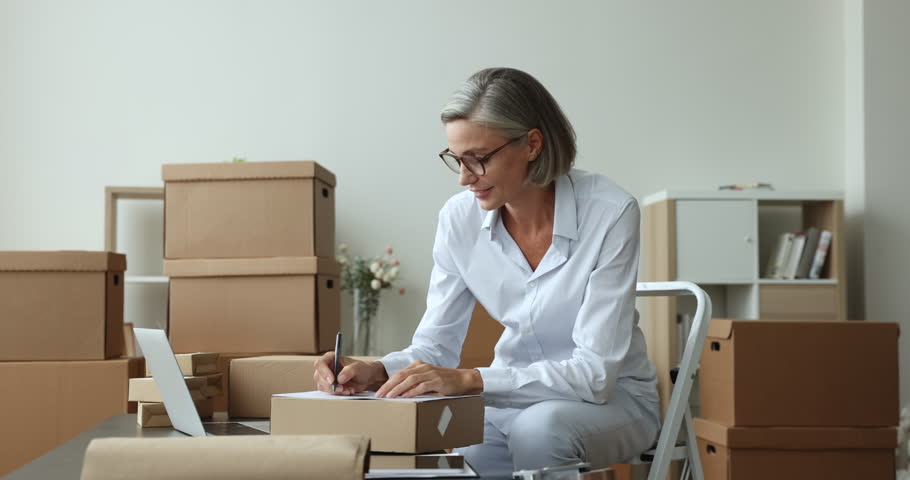 Attractive mature businesswoman, small business owner prepare goods for sending, writes customer data, shipping parcel box using fast and safe transporting services. Shipment, delivery, dropshipping Royalty-Free Stock Footage #1102040397
