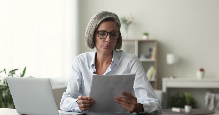 Smiling middle-aged business woman sit at desk in office, looking through paper documents, read great news, got new job, opportunity taxes refund, loan approval, salary increase, career advancement Royalty-Free Stock Footage #1102040433
