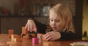 Playful blond girl is busy at the table full of bright orange wooden toy blocks, cute child builds street with houses at kitchen. Kids leisure activity. Nice home interior. High quality 4K footage
