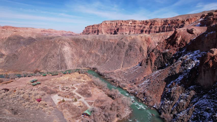 A long river with green water in the Charyn Canyon. There is white snow in places. Bushes and trees grow. The river runs through a canyon among rocks and cliffs. Mountains are visible. Drone view