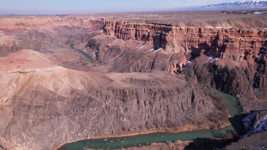 A long river with green water in the Charyn Canyon. There is white snow in places. Bushes and trees grow. The river runs through a canyon among rocks and cliffs. Mountains are visible. Drone view