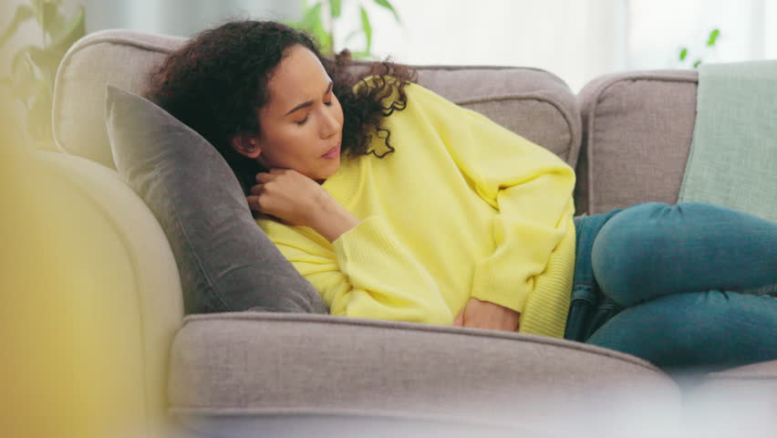 Stomach, pain and woman on a sofa with digestion, problem or suffering from pms. Tummy, virus and sick female with constipation, bloated or endometriosis in living room, unhappy from menstrual cramps | Shutterstock HD Video #1102058847