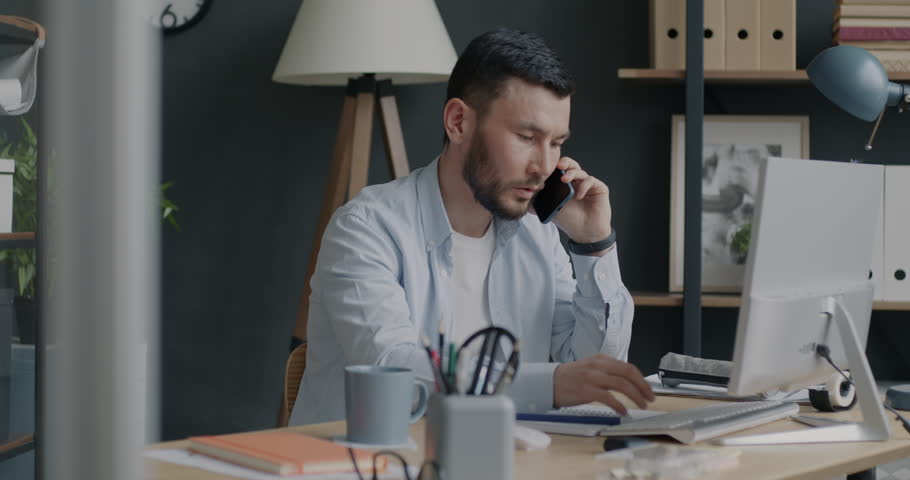 Portrait of young biracial man working with computer and talking on mobile phone in office. Professional conversation and people concept.
