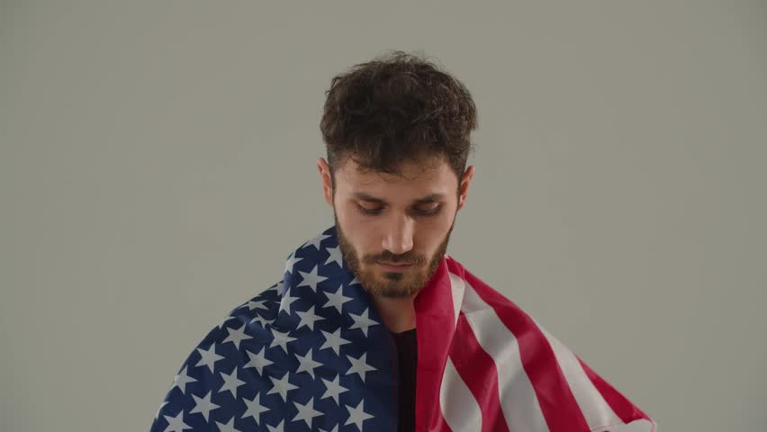 Young curly man covered in the American flag. A serious young man with an American flag is looking at the camera while standing against a gray wall.