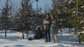 A married couple walks through the forest on a winter sunny day. Two young people are standing in the middle of a snowy forest with a map in their hands and are discussing something.