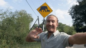 Young man taking selfie on the side of the road near elephants crossing sign in Sri Lanka national park. Man video chatting selfie while enjoying road trip adventures in Asia 