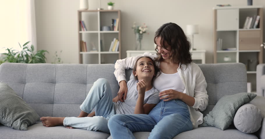 Happy attractive mother talks to little pre-teen daughter seated on sofa at home, family relaxing together, enjoy pleasant friendly conversation, warm communication on leisure. Motherhood, upbringing Royalty-Free Stock Footage #1102066341
