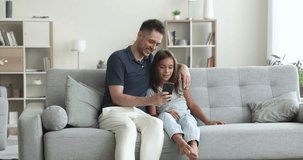 Loving dad teach, explain, show to daughter new mobile app, spend time together with cool, amusing application sit on sofa look at smartphone screen. Young generation use modern gadget, internet usage