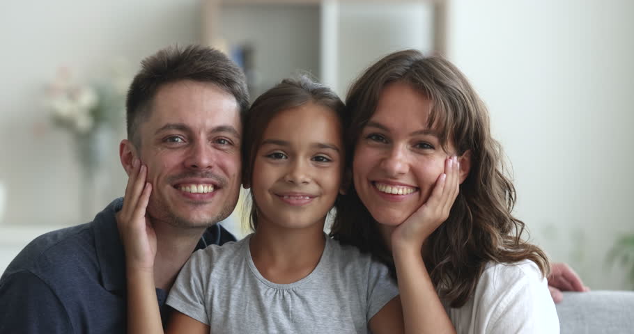 Beautiful Hispanic family close up portrait, parenthood. Happy faces of young woman, man and little adorable daughter touch loving parents cheeks smile look together at camera seated on couch at home Royalty-Free Stock Footage #1102066457
