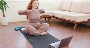 woman with long red hair doing physical exercise watching workout video on laptop, attractive girl following online training lesson, sitting on mat with crossed legs, performing under chin exercise