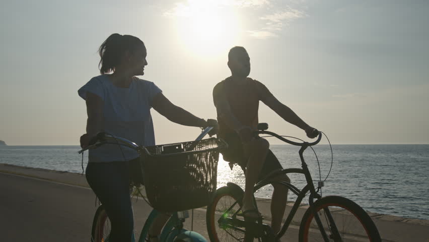 Caucasian couple enjoying a ride on beach cruiser bikes relaxed pedaling and talking near the beautiful sea illuminated by the afternoon sun, handheld shot. Royalty-Free Stock Footage #1102067157