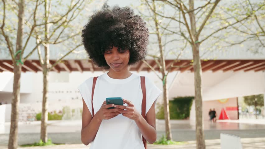 Happy African American student girl with backpack walking outdoors in city and enjoy online communication, scroll social media app on cell phone device on university campus background.