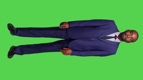 Vertical video: Front view of startup worker talking to people and waving in studio, wearing business suit over full body green screen. Young businessman having conversation on camera, calling someone