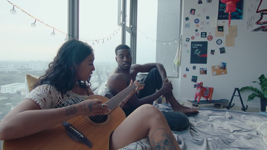 Young couple in love resting on bed in their apartment, girl playing the guitar and singing, boyfriend chilling and listening to her music with pleasure