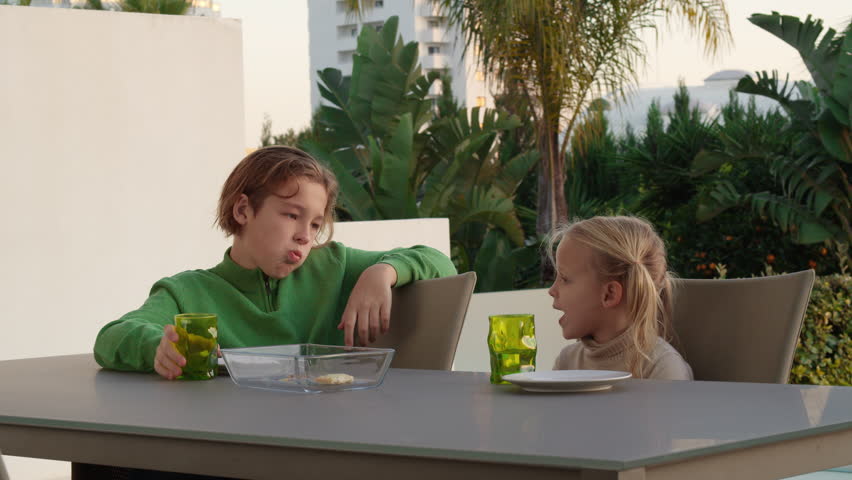 Brother and sister together eating pizza and drinking juice outdoors, with palm trees in the background