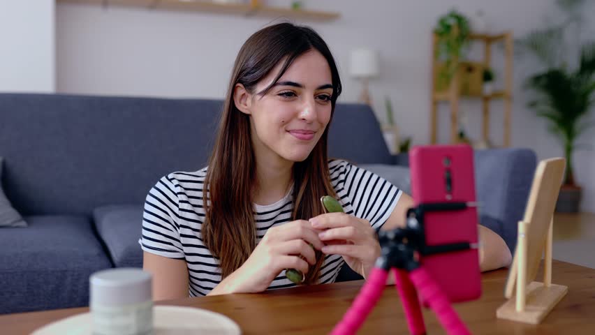 Young influencer woman recording video for social media network massaging facial muscles using natural rose quartz face roller on cheek