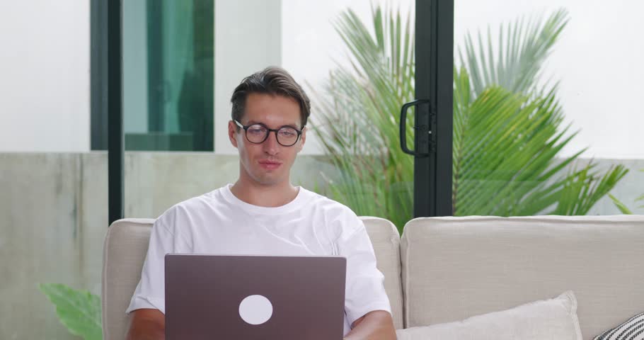 Satisfied and smiling male freelancer works on a laptop while sitting at home on the sofa in a comfortable environment. Millennial man in glasses enjoys using a laptop at home on the sofa