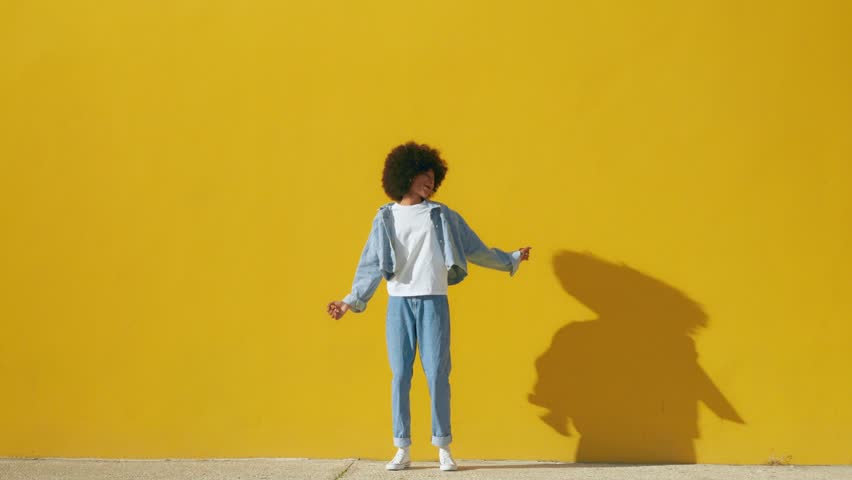 Playful happy young African American woman wear stylish blue denim clothes dancing and jumping funky black teen fashion girl on summer yellow background with copy space. Steady shot, full body | Shutterstock HD Video #1102084097