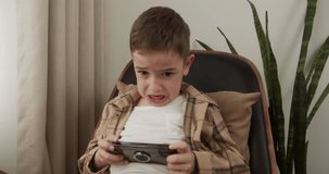 Handsome boy of 6 years old plays a mobile game on a smartphone at home while sitting in an armchair. Adorable kid boy playing mobile phone. Kid using phone for gaming.Child playing video game indoors