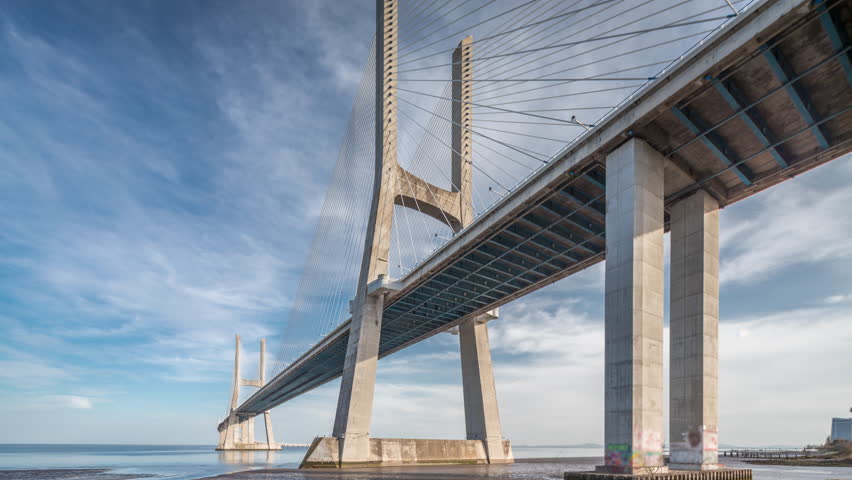 Vasco da Gama bridge timelapse hyperlapse with reflection on water and blue cloudy sky. Cable-stayed bridge and Tagus river. Lisbon, Portugal.