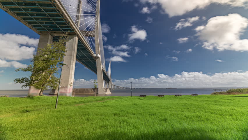Vasco da Gama bridge timelapse hyperlapse with green lawn on a waterfront and blue cloudy sky. Cable-stayed bridge and Tagus river. Lisbon, Portugal.