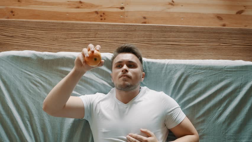 A person is lying on a bed and throws an apple up with his hand and catches it. The guy thinks. Shot from above. 