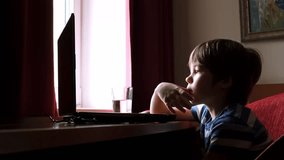 A little boy sits at a table carefully is looking at laptop screen. The child take finger in mouth. Watching cartoons. Attention. Concept of infection through dirty hands. Nervous kid. 4k. Technology.