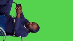 Vertical video: Side view of businessman using laptop and listening to music in studio, wheelchair user with impairment. Young man with chronic disability working on computer, enjoying song on headset