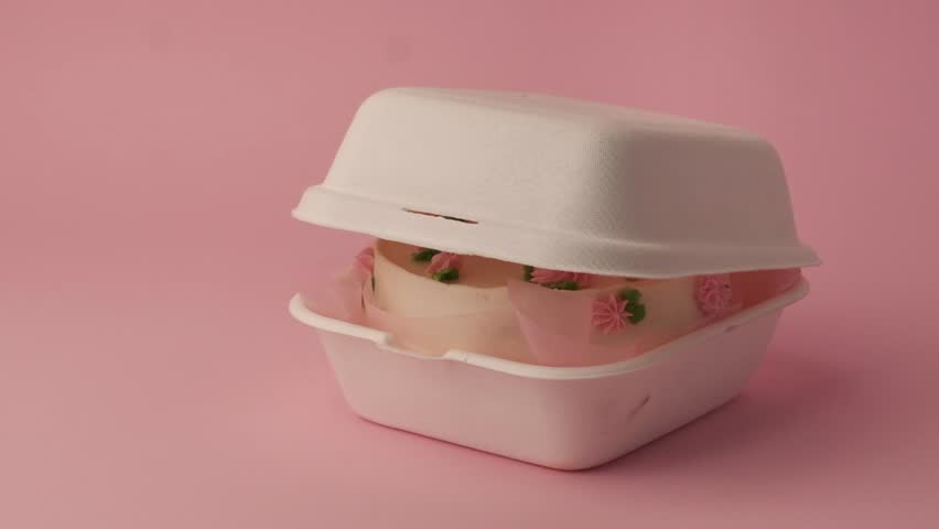 Small traditional Korean style bento cake decorated with pink whipped cream flowers. Woman hand opens white gift box with little spring cake on the pink pastel background