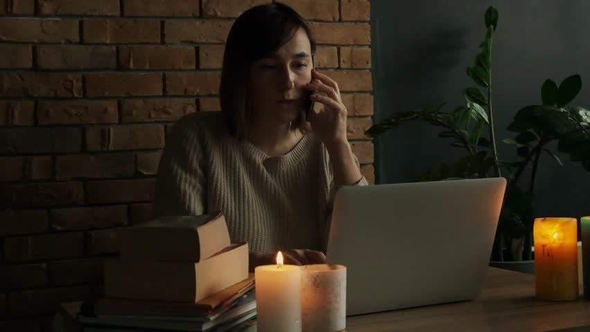 Amazed caucasian woman using laptop to read the news via internet at home during electricity outage or blackout with lit candles. Woman shocked by the bad news while talking on the phone Royalty-Free Stock Footage #1102114619