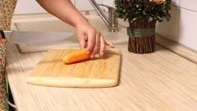 Video of cropped female hands cutting, slicing and chopping fresh organic vegetable carrot on wooden cutting board on table in domestic kitchen. Farm ingredients for healthy salad or soup, 4k