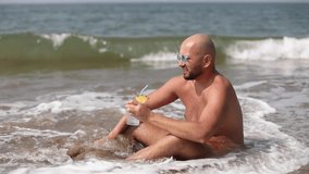 Satisfied, relaxed bald unshaven man in sunglasses and shorts drinking cocktail with straw and lemon, sunbathing in sea waves. Joying tide, splashing water. Island resort. Video, animation