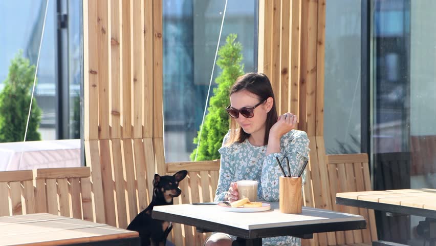 Young woman sits in street cafe, with little dog, has breakfast. Girl treats lapdog with her breakfast.