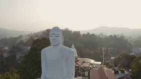 Drone point of view of big white sitting Buddha overlooking the green lush hills around. Aerial view of buddhist temple in Kandy, Sri Lanka at sunset