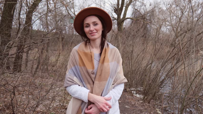 A young woman in a hat and white sweater walks near dry plants. Slow motion portrait of a woman in a hat with a scarf. Beautiful young woman in her thirties