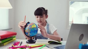 Happy Asian child girl student sitting using laptop computer to online study with search the map and country on the model globe in geography lesson in the room at home. Homeschool education concept. 