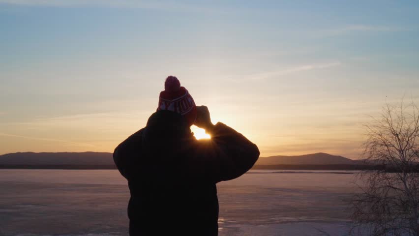 A young man looks through binoculars at a frozen lake and mountains at sunset. The rays of the bright sun gild everything around. Travel and tourism.