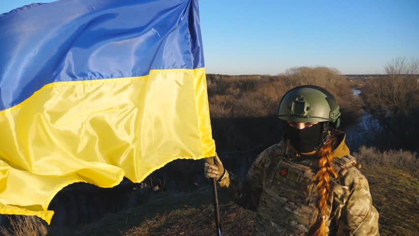 Young girl in camouflage uniform holds a waving flag of Ukraine. Female military medic of ukrainian army lifting blue-yellow banner at countryside. Victory against russian aggression. End of war Royalty-Free Stock Footage #1102132585