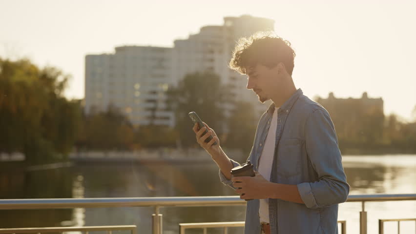 Side View of the Young Man Walking on the Street While Sunset, Using his Smartphone, Texting With Friends. Caucasian Male Outside Scrolling Mobile While Going