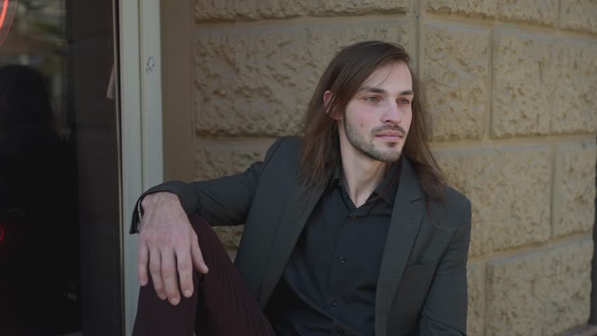 Portrait of a relaxed handsome young man smiling and looking at camera in a urban street. man sits close to window. black shirt and jacket