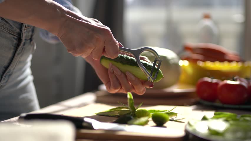 Housewife peeling off cucumber with a peeler tool in close up. Female person peels vegetables for lunch at home