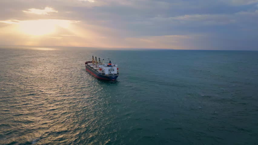 Drone view bulk carrier ship sailing to picturesque sunset. Big cargo vessel floating dark deep ocean at summer evening. Bright golden sunlight shining reflecting in sea surface. Beauty marine sundown