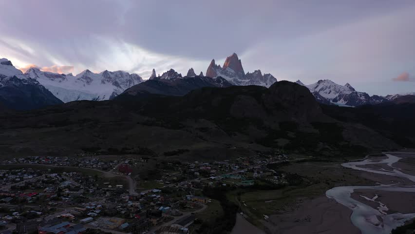 Traveler Man with Orange Backpack Standing and Looking at Mount Fitz Roy. El Chalten Town. Hills and Snow-Capped Mountains. Andes, Patagonia, Argentina. Aerial View. Drone Goes Backwards at Low Level