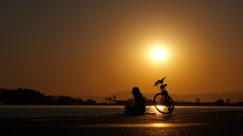 Lonely female with a bicycle at sunset. Silhouette of a female cyclist resting near the sea bay at a beautiful sunset. Royalty-Free Stock Footage #1102165415